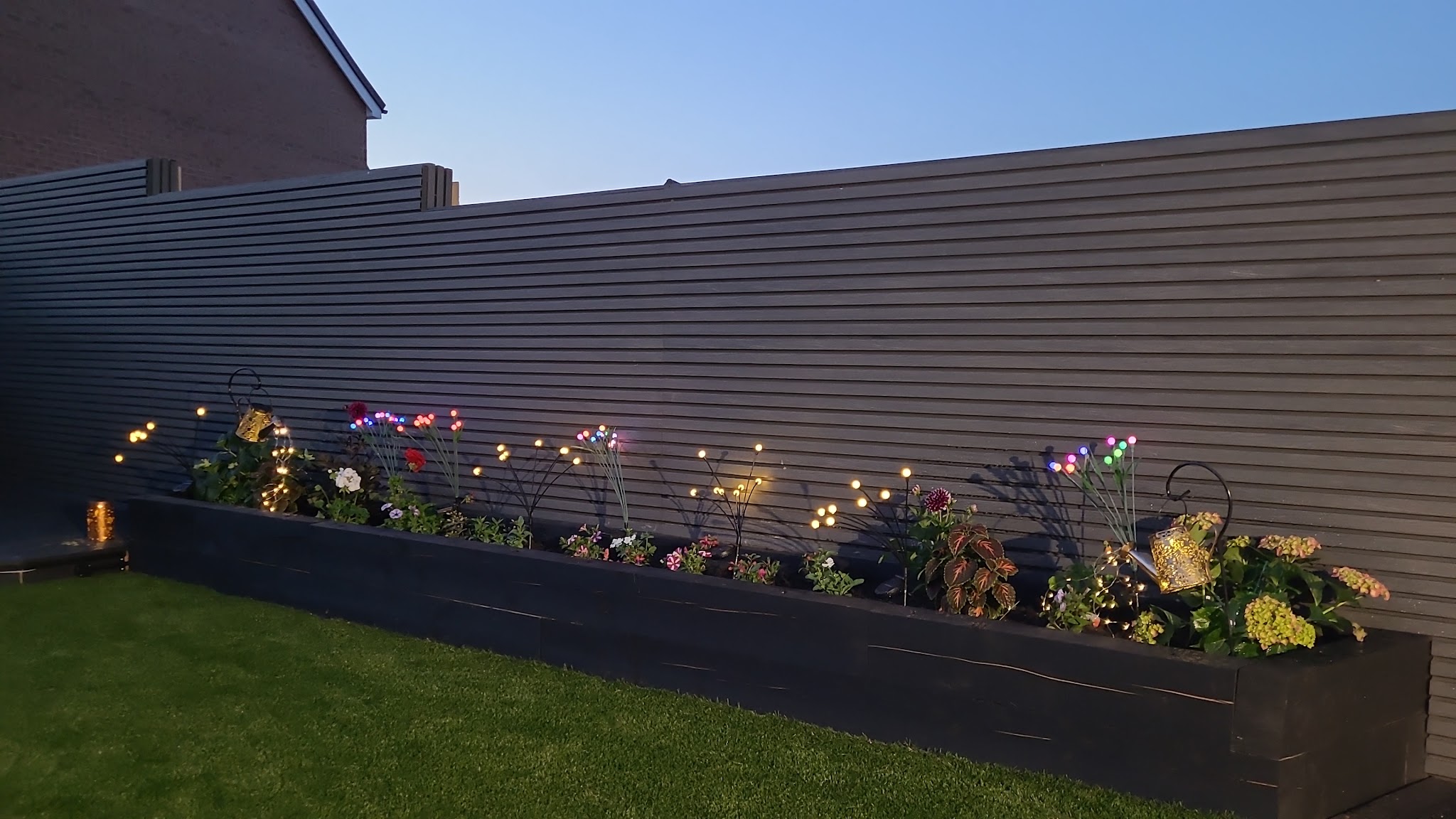 Long raised planter bed lit up at dusk beside an artificial lawn